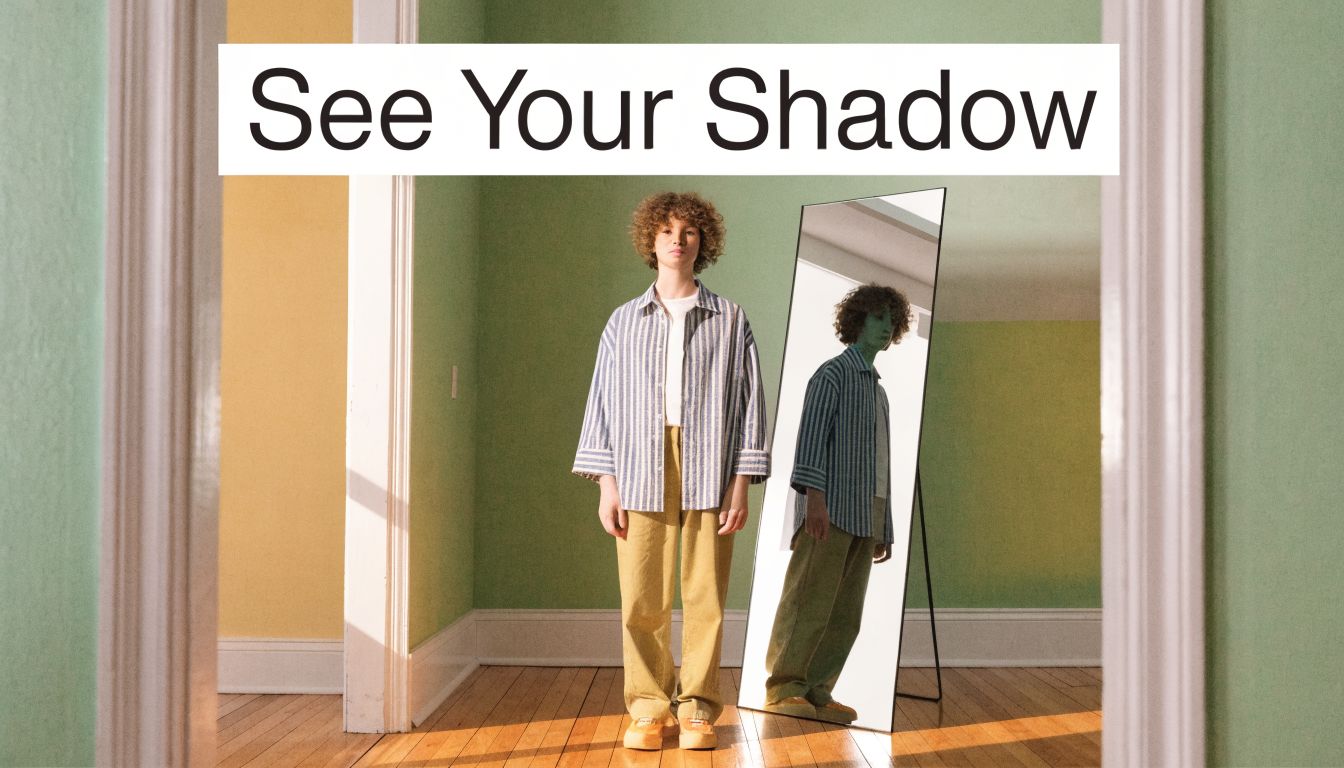 A person with curly hair standing in front of a floor mirror inside a minimalist room.
