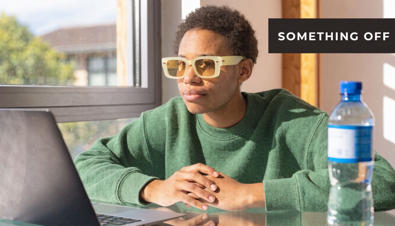 A person wearing yellow-tinted sunglasses sitting at a desk with a laptop and a water bottle.